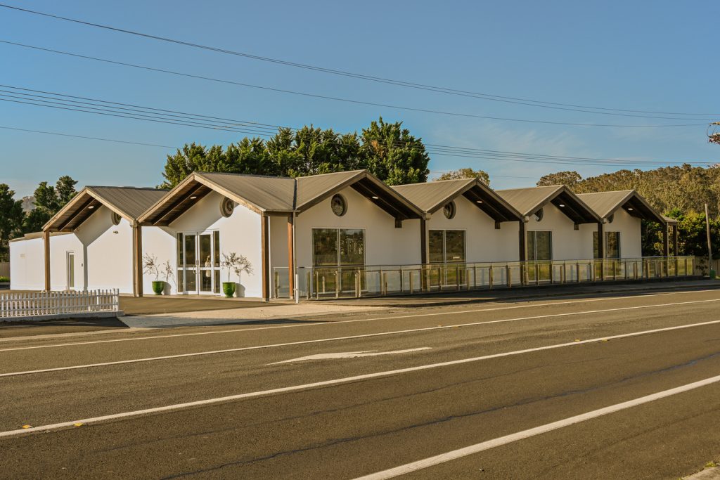 A modern, white single-storey building with multiple pitched roofs, large glass windows, and wooden accents sits beside a road on a sunny day. There are trees behind the building and clear blue sky overhead. by H Parsons Funeral Directors