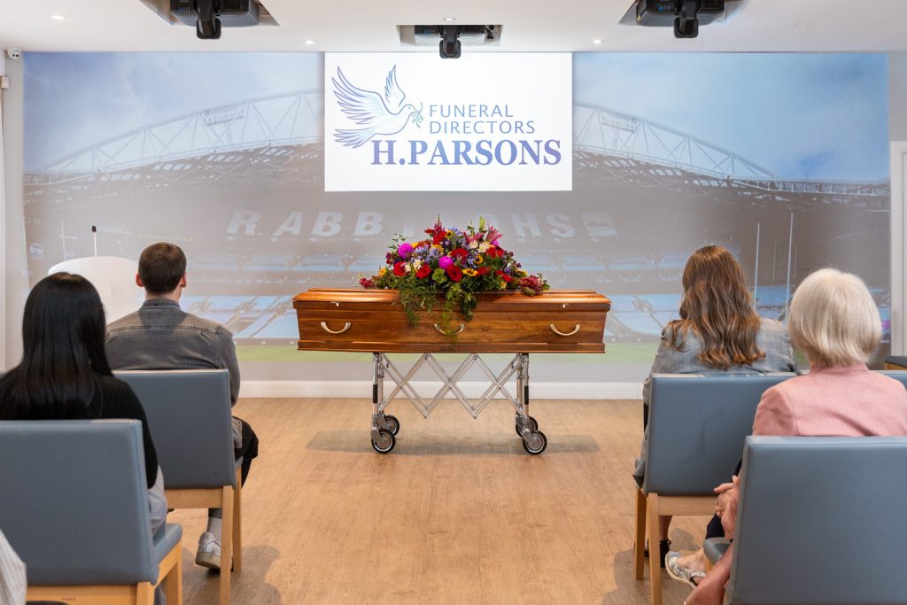 A group of people sit facing a wooden coffin adorned with flowers in a modern funeral chapel. The wall behind displays "FUNERAL DIRECTORS H. PARSONS" alongside a dove, stadium image, and Services Directory details. by H Parsons Funeral Directors