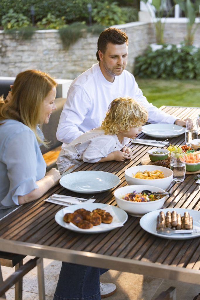 A family of three, two adults and a child, sits at an outdoor table set with various dishes, including grilled food, crisps, and salad, enjoying a meal together in a garden setting. by H Parsons Funeral Directors