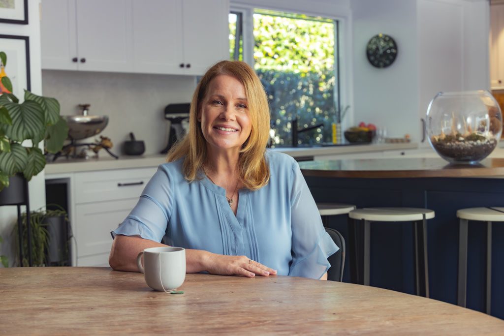 A woman with light brown hair and a blue blouse sits at a wooden table, smiling, with a white mug in front of her in a bright, modern kitchen. by H Parsons Funeral Directors