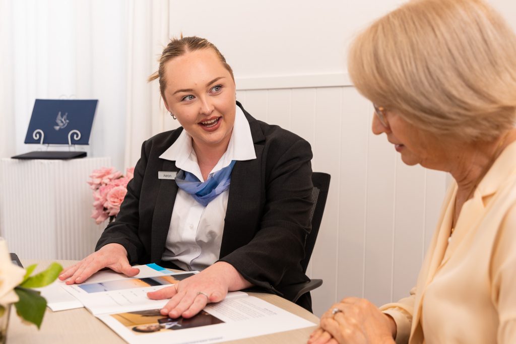 A woman in a suit explains documents about funeral costs to an older woman at a desk. Both are seated, with the adviser smiling and gesturing to printed papers. Pink flowers and a blue folder are visible in the background. by H Parsons Funeral Directors