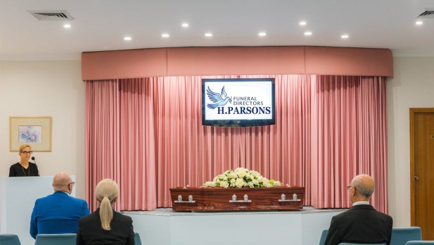 A small funeral service with four attendees seated, a coffin adorned with white flowers at the front, and a woman standing at a lectern. A screen above displays "Funeral Directors H. Parsons. by H Parsons Funeral Directors