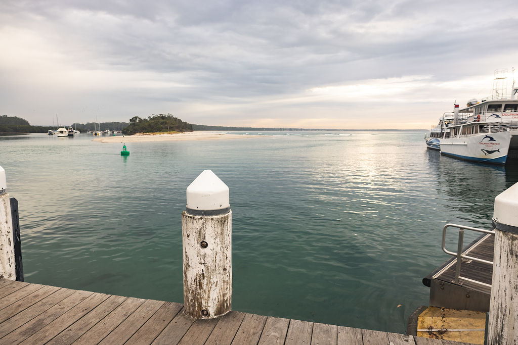 A calm waterfront scene with a wooden pier, boats moored to the right, and a small island in the distance under a cloudy sky—a peaceful spot to reflect on what to do if someone dies abroad or when considering repatriation death procedures. by H Parsons Funeral Directors