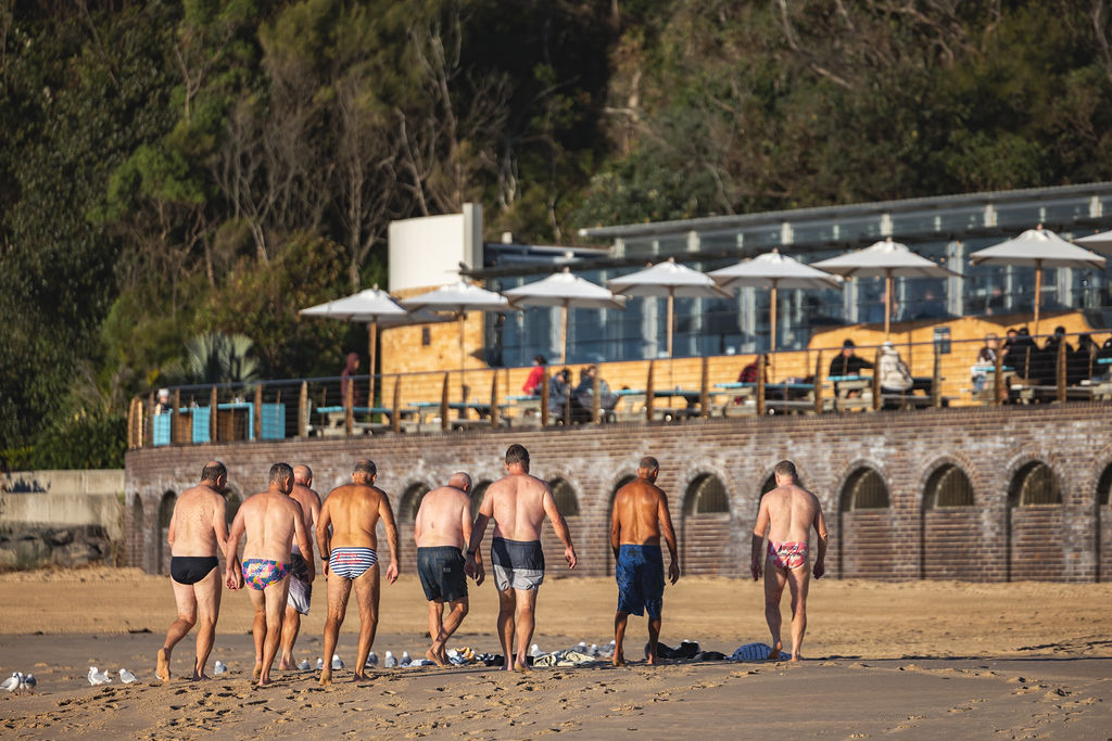 A group of older men in swimming trunks walk together on a sandy beach towards the water, perhaps gathering to arrange a funeral or simply reminiscing, as a beachside café and people under umbrellas rest in the background. Trees line the area behind the café. by H Parsons Funeral Directors