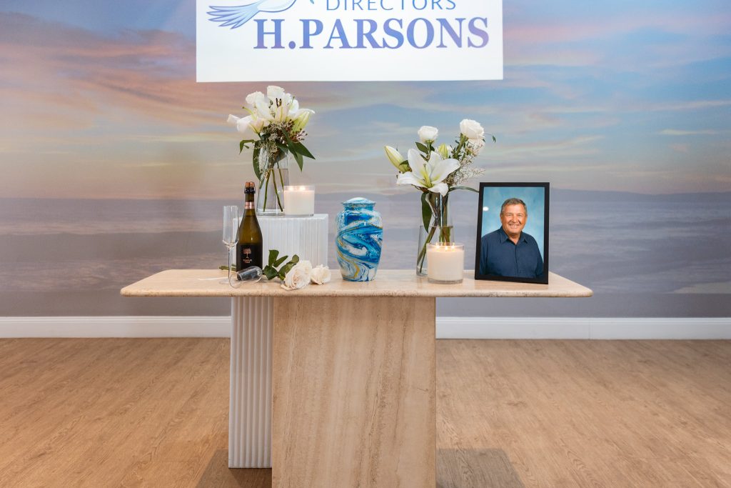 A memorial table with flowers, candles, a blue urn, champagne, seashells, and a framed photo of an older man is set beneath a sign reading "H. Parsons" against a serene ocean sunset—a heartfelt way to arrange a funeral by the sea. by H Parsons Funeral Directors