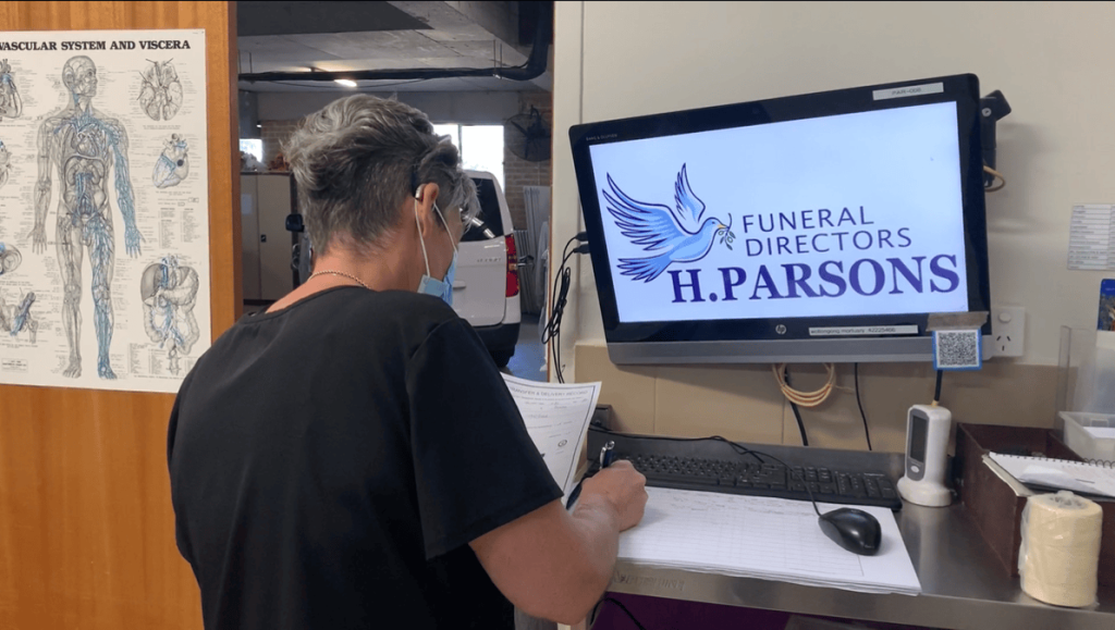 A person with short hair and a mask writes on a clipboard at a desk, facing a computer monitor displaying the H. Parsons Funeral Directors logo. A medical anatomy poster is on the wall nearby. by H Parsons Funeral Directors