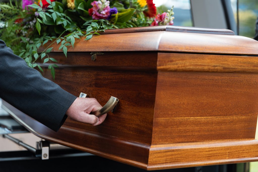 A person in a dark suit holds the handle of a polished wooden coffin adorned with a colourful flower arrangement, featured in the Services Directory. by H Parsons Funeral Directors