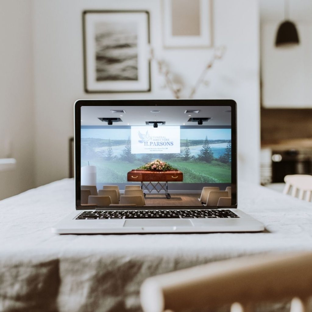 A laptop displays a virtual funeral service with a flower-adorned coffin and chairs, set on a table in a cosy, blurred home environment, reminiscent of Parsons Ladies' thoughtful attention to life's important moments. by H Parsons Funeral Directors