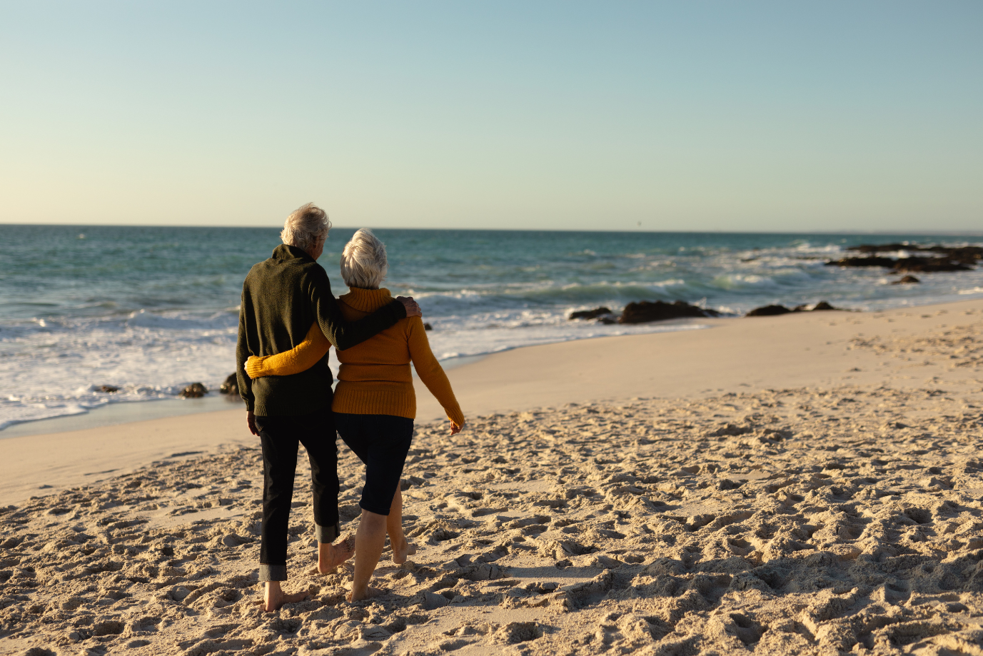 An older couple walk barefoot on a sandy beach, arm in arm, facing the sea under a clear sky. The beach is mostly empty with waves gently rolling onto the shore. by H Parsons Funeral Directors