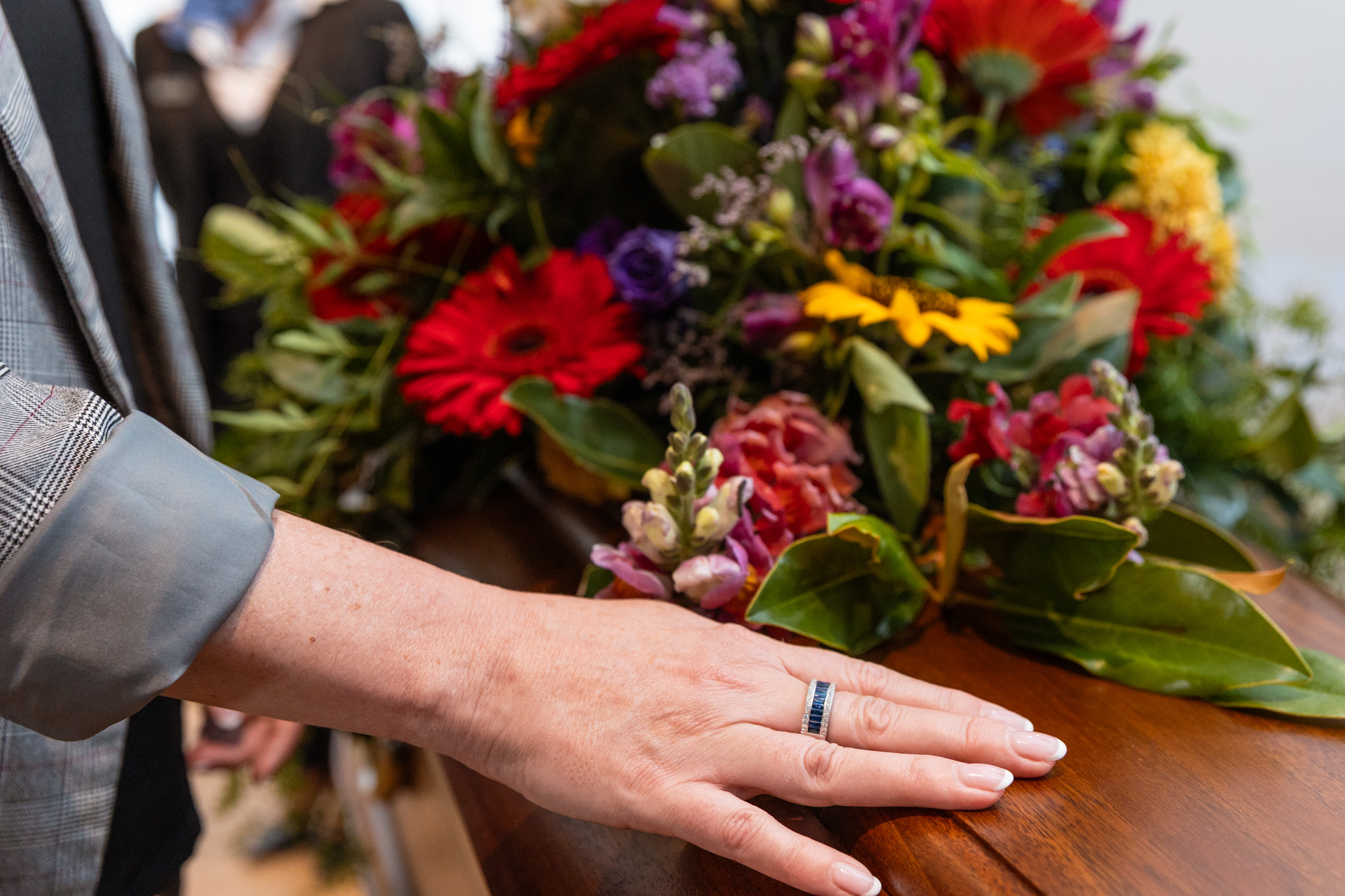 A person rests their hand on a wooden coffin adorned with a colourful flower arrangement, suggesting a funeral or memorial service, reflecting the spirit of Dying to Know Day—a time to open conversations about death, dying, and end-of-life choices. by H Parsons Funeral Directors