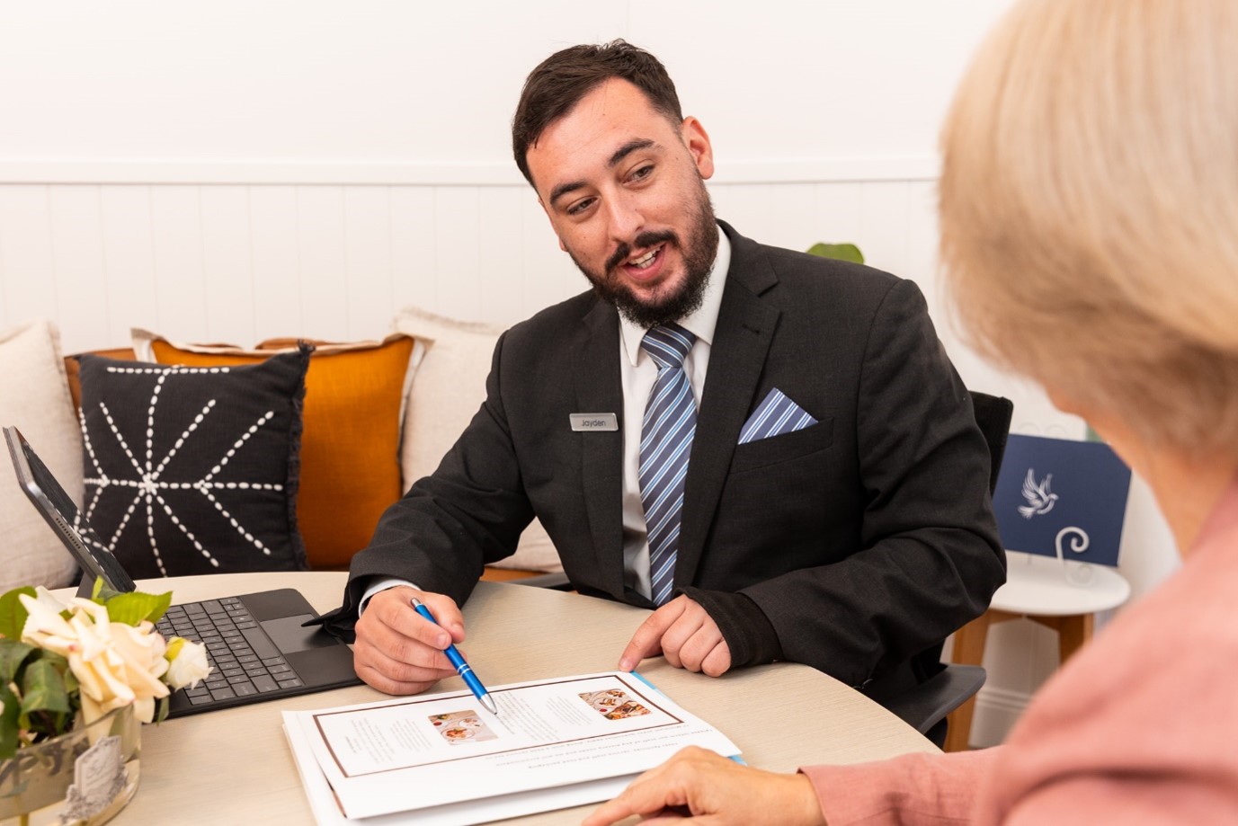 A man in a suit gestures to a document whilst talking to a woman at a table, perhaps engaging in an important Dying to Know Day discussion. Nearby, a laptop, flowers, and decorative cushions add warmth to the setting. by H Parsons Funeral Directors