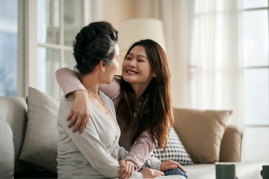 A young woman and an older woman sit on a sofa, embracing and smiling at each other warmly in a bright, cosy living room—a heartfelt moment of connection that beautifully embodies the spirit of Dying to Know Day. by H Parsons Funeral Directors
