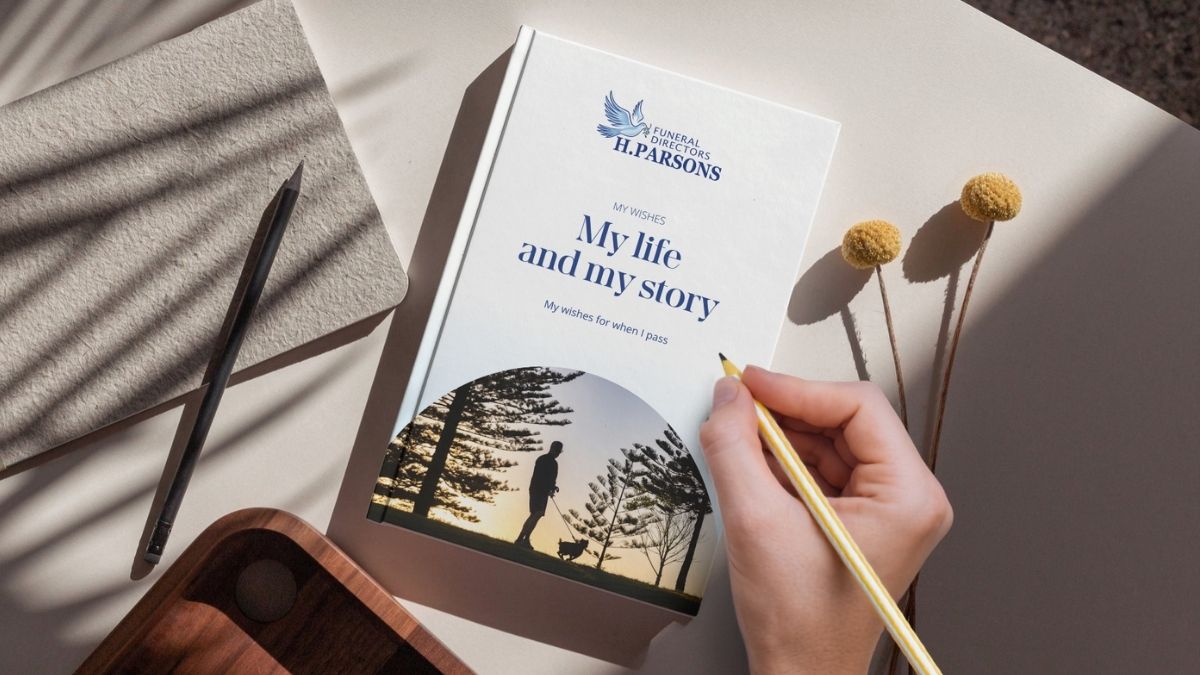 A hand holding a pencil writes in a diary titled “My life and my story.” The cover shows a silhouette of a person and dog with trees. The diary rests on a beige table with an exercise book, tray, and yellow flowers. by H Parsons Funeral Directors