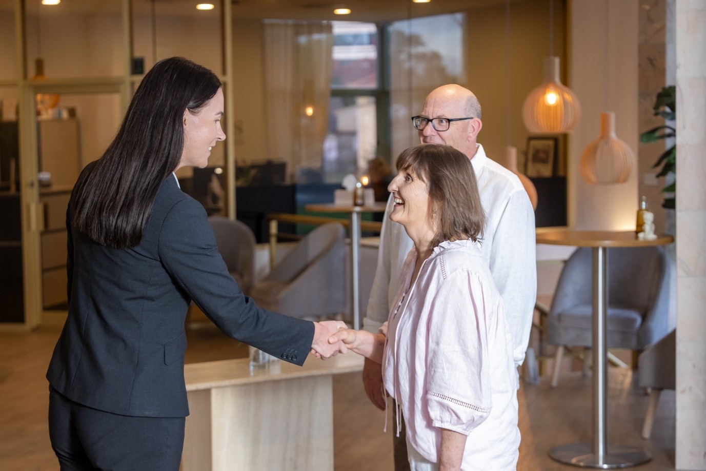A woman in a suit shakes hands with an older woman while an older man stands beside them, smiling, in a modern office lobby with soft lighting and contemporary decor—an atmosphere of thoughtful connection that perfectly reflects the spirit of Dying to Know Day. by H Parsons Funeral Directors