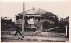 A person walks past an old, weathered house with a tin roof, a verandah, and overgrown plants. A wooden pole stands in front, and the pavement is cracked. The image is black and white. by H Parsons Funeral Directors