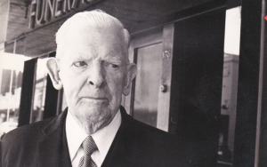 An elderly man in a suit and tie stands outside a building with a “FUNERAL” sign visible above the entrance. The image is in black and white. by H Parsons Funeral Directors