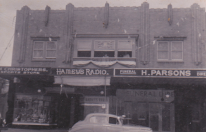 A vintage black and white photo shows shopfronts for sports, radio, and funeral businesses, with an old-fashioned car parked in front of the building. by H Parsons Funeral Directors