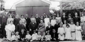 Black and white photo of a large family, with adults standing in rows, and children seated in front, posing outdoors in front of a wooden building. by H Parsons Funeral Directors