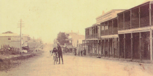 Historic sepia photo of a dusty high street lined with wooden buildings; a man and child stand beside a bicycle in the foreground, with a few people in the distance and a sign reading “H. Parsons Wholesale Grocer”. by H Parsons Funeral Directors