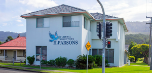 A two-storey white building with a sign reading “Funeral Directors H. Parsons” and an illustrated bird. There is a traffic light, a “DIP” road sign, and a nearby single-storey house. Trees and hills are in the background. by H Parsons Funeral Directors