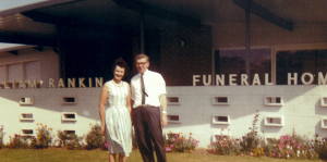 A woman in a light dress and a man in a shirt and tie stand smiling in front of a building with “FUNERAL HOME” on the sign. Flowers line the building’s front. by H Parsons Funeral Directors