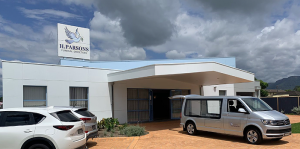A modern white building with a sign reading “H. Parsons Funeral Directors.” Two white vehicles, one a hearse, are parked outside under a cloudy sky. by H Parsons Funeral Directors