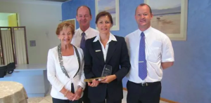 Four adults in business attire stand indoors, posing and smiling. The woman in the centre holds a glass award. There are framed pictures on a blue wall behind them. by H Parsons Funeral Directors