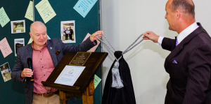 Two men unveil a plaque at an indoor ceremony; one holds a microphone, the other pulls a cord. Photos and documents are displayed on a board behind them. by H Parsons Funeral Directors