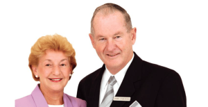 An older woman in a lavender blazer and an older man in a black suit with a name badge stand smiling together against a white background. by H Parsons Funeral Directors