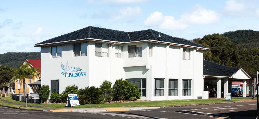 A white, two-storey building with a sign reading “Funeral Directors H. Parsons” on the side, located on a corner plot with neatly trimmed bushes and a small lawn. by H Parsons Funeral Directors