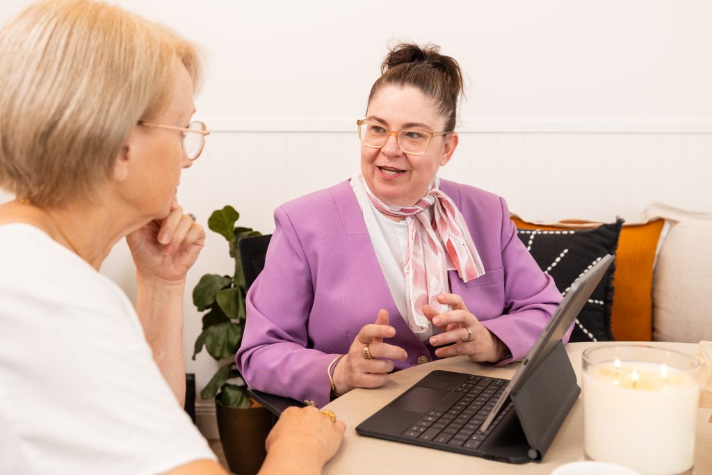 Two women sitting at a table with a laptop, one gesturing while talking to the other about understanding grief. by H Parsons Funeral Directors