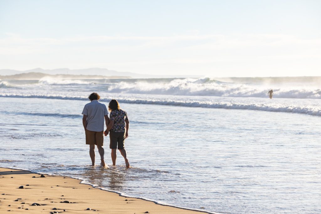 A couple walks along the shore holding hands, seeking an understanding of grief together. by H Parsons Funeral Directors